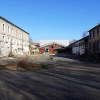 Cour d’une ancienne friche industrielle entre deux bâtiments dégradés sous un ciel bleu.